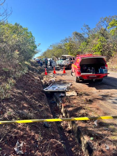 Acidente de trânsito em uma estrada com veículos danificados e presença de equipes de emergência. Caminhão vermelho com equipamentos ao lado.
