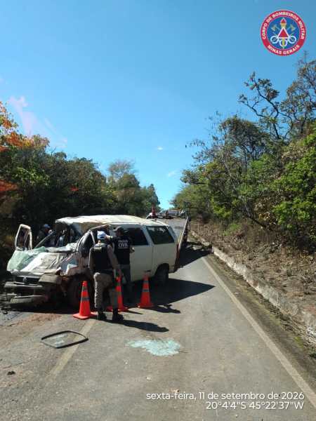 Imagem de um veículo acidentado na estrada, com cones de sinalização e pessoas trabalhando no local. A vegetação está ao redor da pista.
