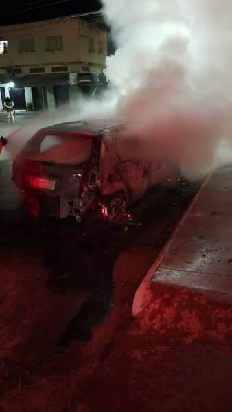 Carro em chamas com fumaça saindo, em um ambiente urbano à noite.