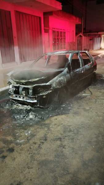 Carro queimado abandonado na rua, com fumaça saindo e iluminação avermelhada ao fundo.