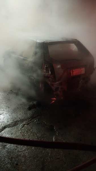 Carro danificado coberto por fumaça em uma área com chão de asfalto.