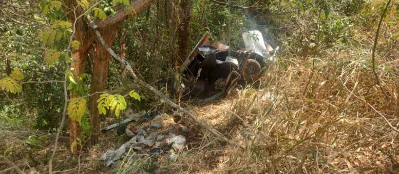 Um carro acidentado em uma área de mata, cercado por vegetação densa e detritos.