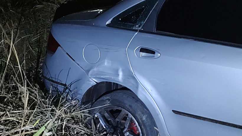 Um carro prateado danificado parado em um terreno gramado à noite, com o painel traseiro amassado e a roda visível.