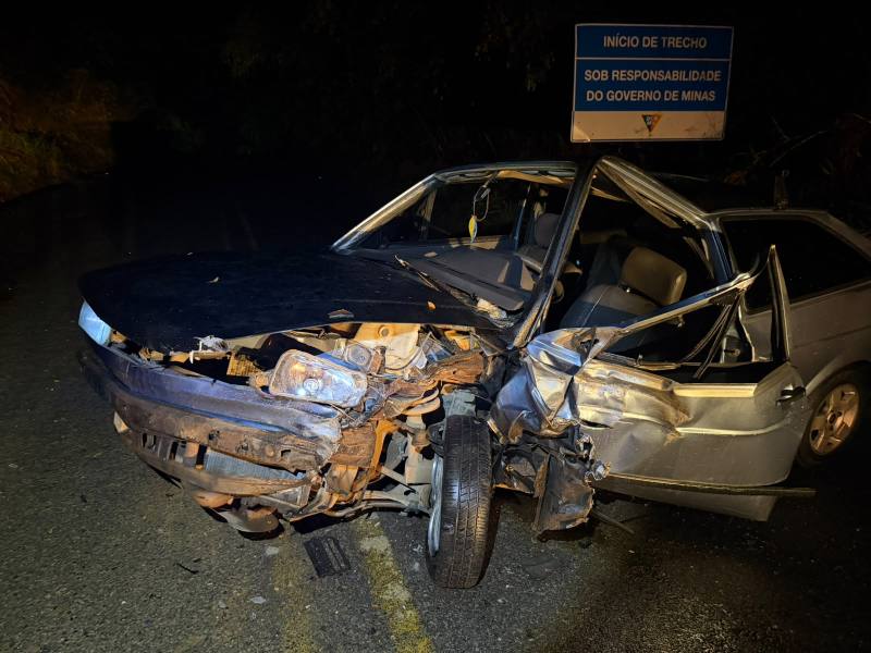 Carro colidido em uma estrada, com a frente parcialmente destruída e a porta do lado do motorista aberta. Placa indicando o início de trecho sob responsabilidade do governo de Minas.