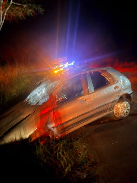 Veículo acidentado em uma estrada durante a noite, com luzes de emergência ao fundo.