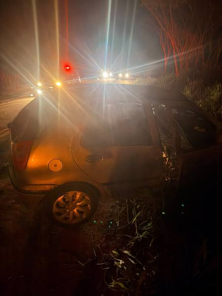 Um carro acidentado à noite, com uma porta aberta e luzes de viaturas ao fundo, emitindo feixes de luz no caminho.