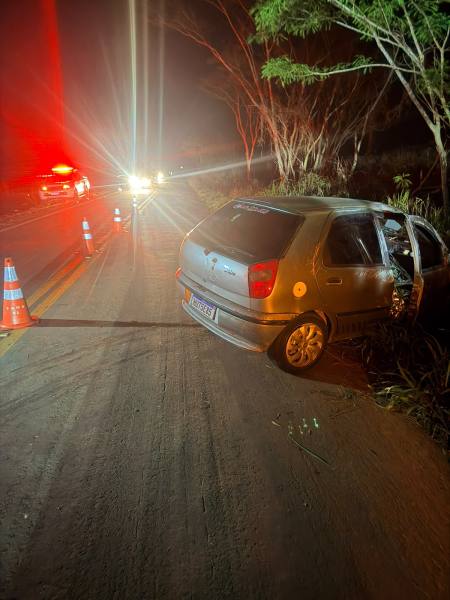 Carro acidentado à beira da estrada à noite, com luzes de emergência visíveis e cones de sinalização ao redor.