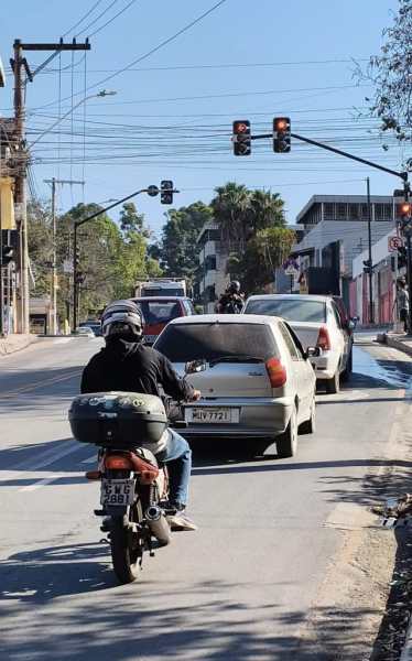 Motociclista aguardando no semáforo, com carros parados à frente e árvores nas laterais da rua.
