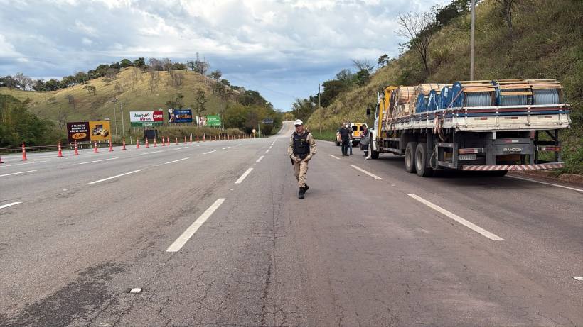 Policial caminhando em rodovia com caminhão ao fundo e placas de publicidade ao lado.