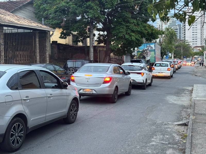 Imagem de um trânsito intenso em uma rua urbana, com vários carros estacionados e em movimento, cercados por edifícios e árvores.