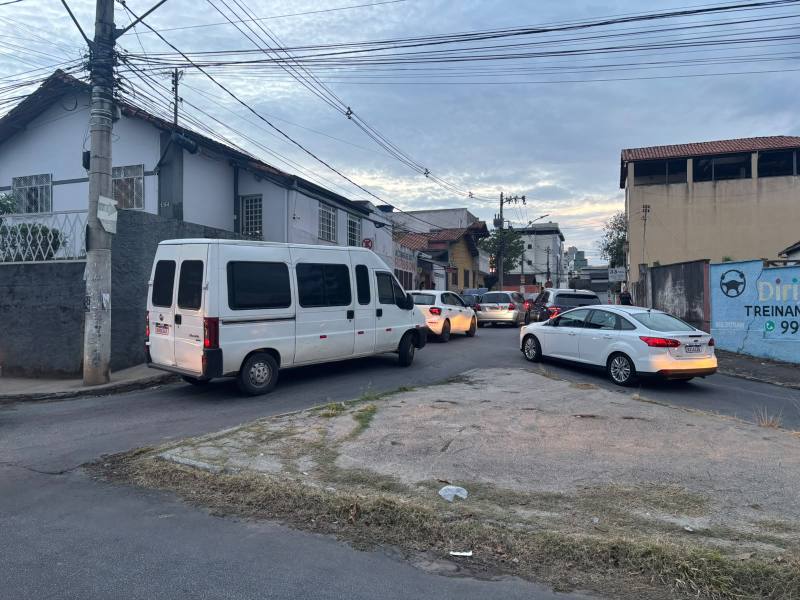 Tráfego intenso em uma rua residencial, com veículos parados e um furgão branco na esquina.