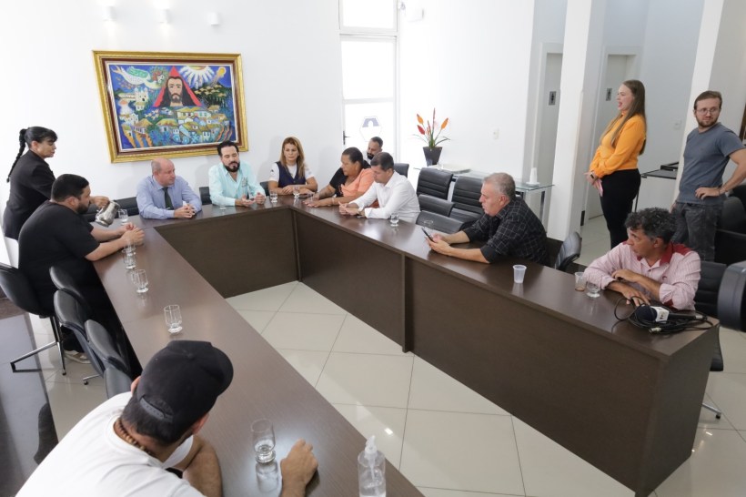 Reunião em sala de conferências com várias pessoas ao redor de uma mesa longa, discutindo assuntos importantes, com uma obra de arte colorida na parede.