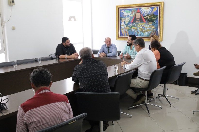 Reunião em uma sala com várias pessoas sentadas ao redor de uma mesa, discutindo assuntos diversos. Uma pintura colorida na parede ao fundo.