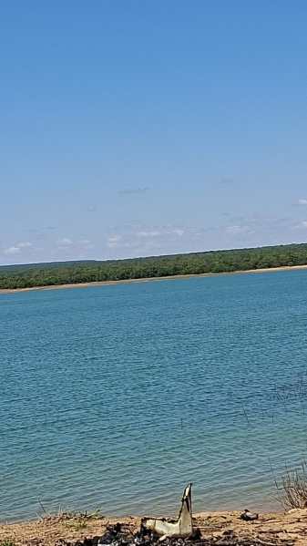 Vista de um corpo d'água azul sob um céu claro, com vegetação verde ao fundo.