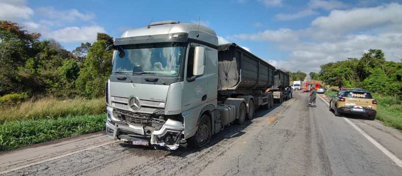 Caminhão prateado danificado parado na estrada, com veículos atrás e vegetação ao fundo.