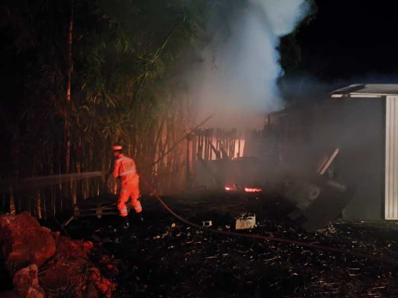 Bombeiro combatendo incêndio em área escura, com fumaça densa e chamas visíveis ao fundo, cercado por vegetação.
