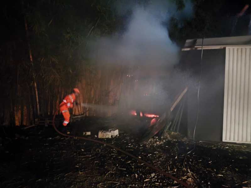 Bombeiro apagando um incêndio em área externa, com fumaça visível e fogo ao fundo.