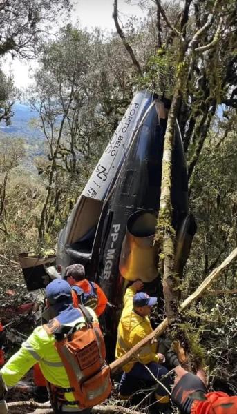 Equipe de resgate trabalhando ao redor de um helicóptero acidentado em uma área arborizada.