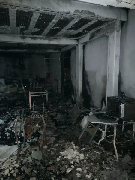 Ambiente interno danificado por incêndio, com móveis queimados eRuínas de paredes e piso cobertos de cinzas e detritos.