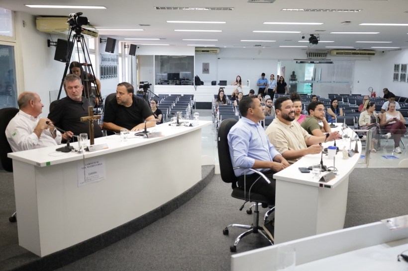 Reunião em um auditório com várias pessoas, incluindo apresentações em mesas, câmeras e uma assistência parcialmente cheia.