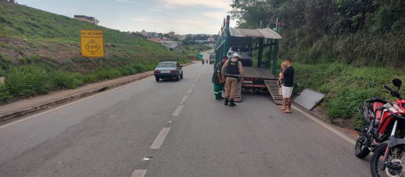 Cena de uma estrada com um veículo de transporte parado e policiais conversando com um motorista, enquanto uma pessoa aguarda ao lado. Placa de advertência visível na pista.