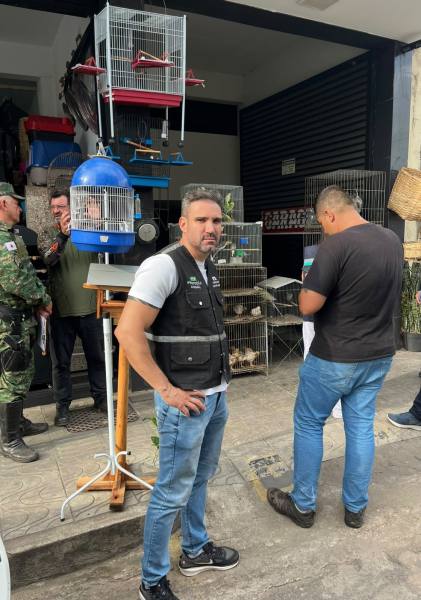 Homem em frente a uma loja com aviários, usando colete, enquanto outros homens estão presentes.