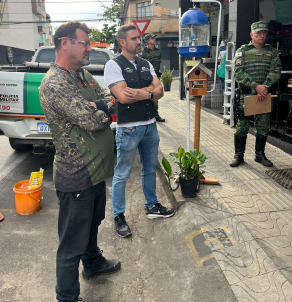 Três homens em uma rua observam um evento, com um veículo da Polícia Militar ao fundo. Um deles está vestindo uma camisa camuflada, outro usa um colete, e o terceiro está em uniforme militar. Um pequeno viveiro de pássaros está visível ao centro.