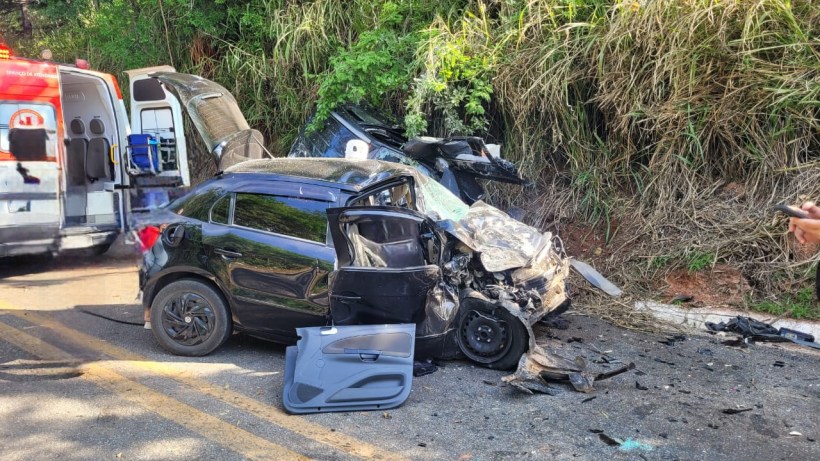Acidente de carro com um veículo preto colidido e danificado em uma estrada, com uma ambulância ao fundo.