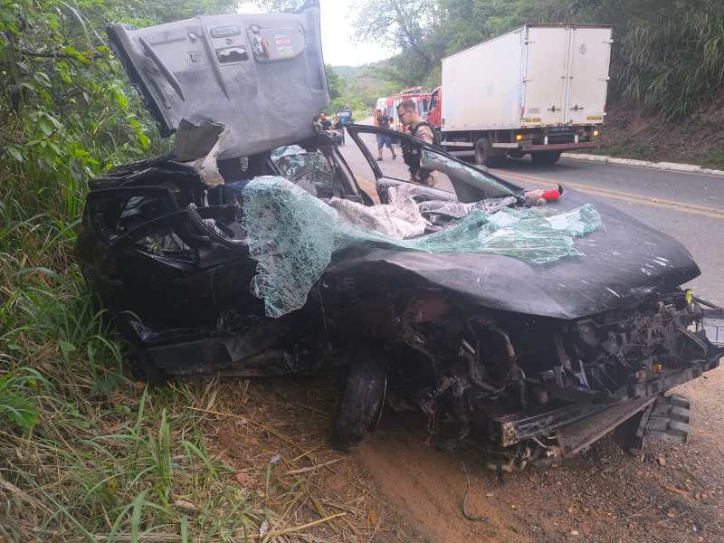 Veículo acidentado na beira da estrada, com carroceria danificada e vidro quebrado, cercado por vegetação.