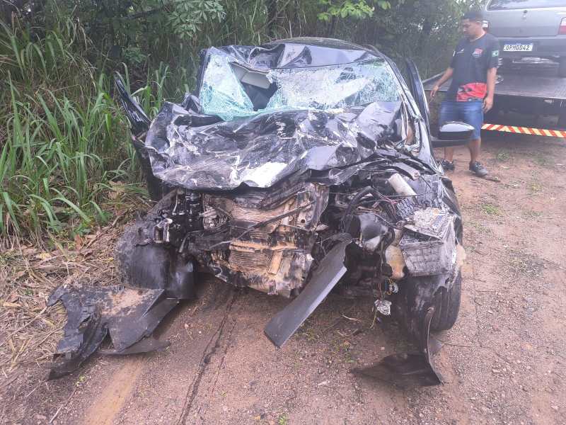 Veículo danificado gravemente em um acidente, com a frente destruída e vidro quebrado, ao lado de uma pessoa em uma estrada rural.
