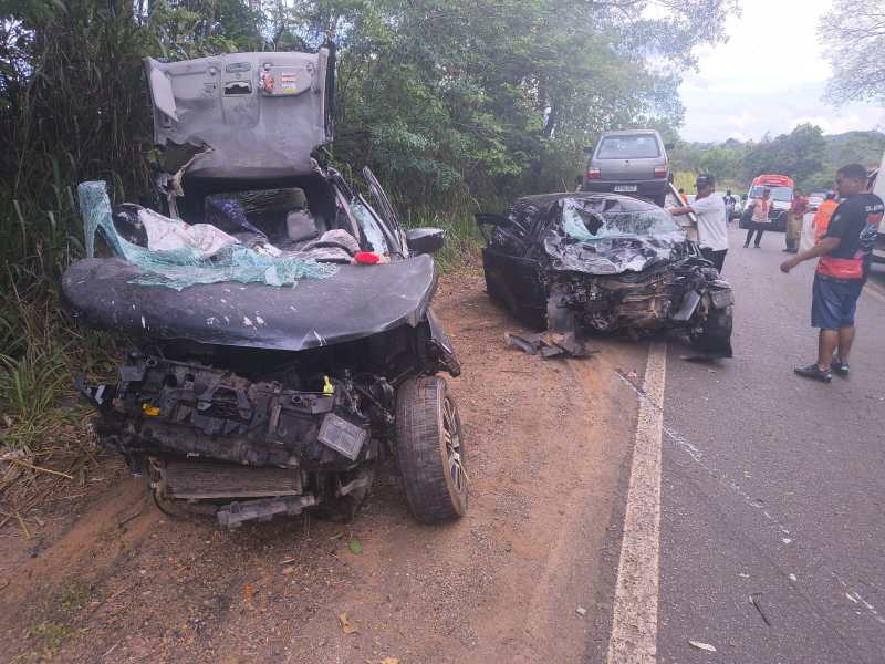 Acidente de carro com dois veículos danificados na estrada, um carro está com o capô aberto e o outro tem a frente destruída, com rodeio de pessoas possivelmente auxiliando na situação.