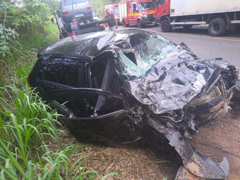Carro preto danificado após acidente, com vidros quebrados e capô amassado, localizado na beira da estrada com vegetação ao redor.