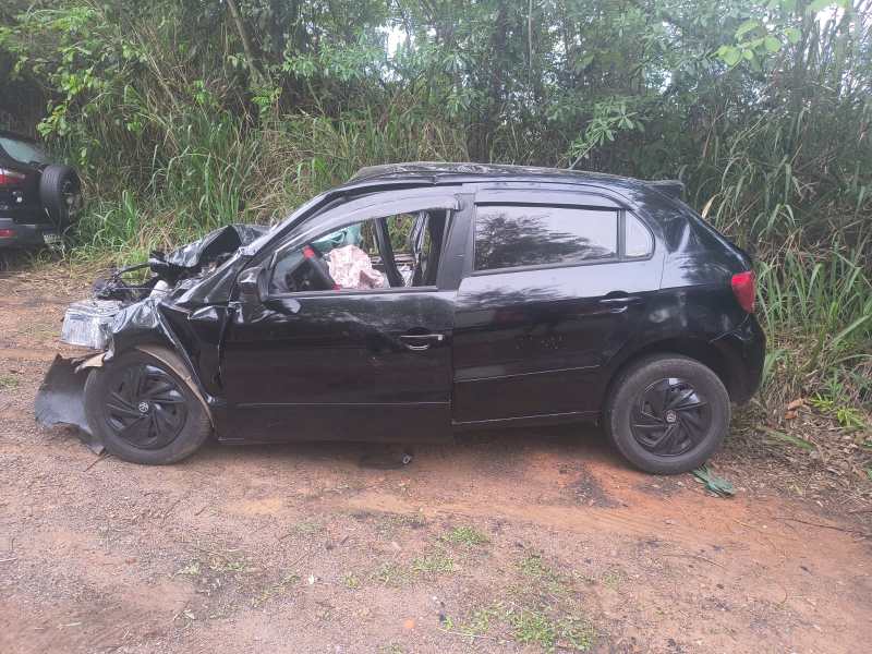 Um carro preto danificado estacionado em uma área cercada por vegetação. O lado esquerdo do veículo apresenta danos significativos, enquanto a porta do motorista está aberta.