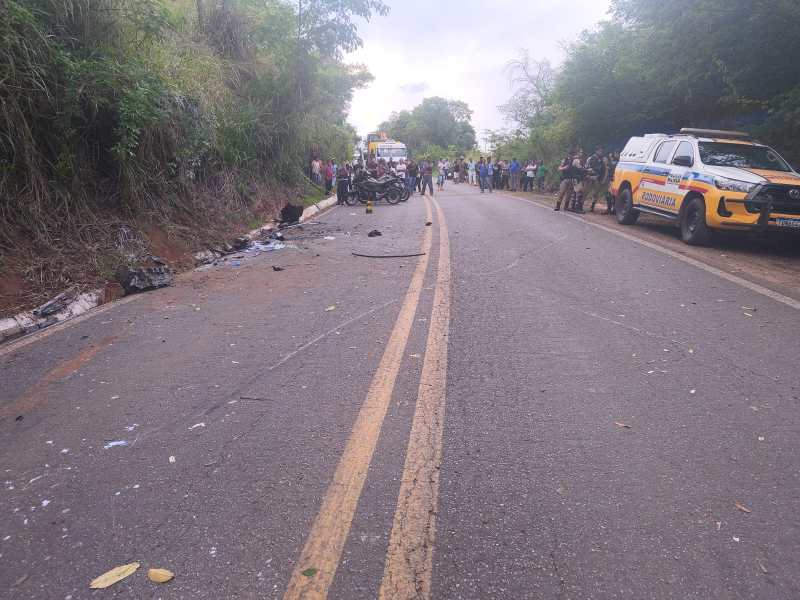 Cena de um acidente de trânsito em uma estrada, com destroços visíveis e vários espectadores ao fundo. Veículos de emergência estão estacionados ao lado da estrada.