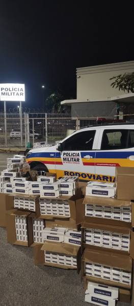 Viatura da Polícia Militar rodoviária ao lado de caixas empilhadas com produtos apreendidos à noite, com um letreiro da Polícia Militar ao fundo.