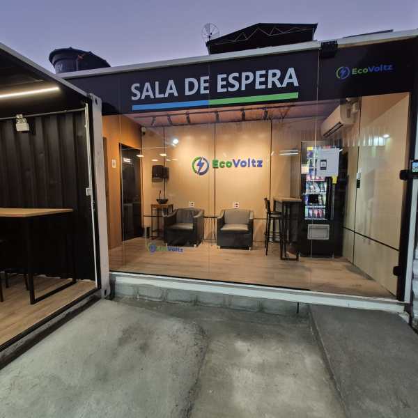 Fachada da sala de espera da EcoVoltz com janelas de vidro, mesas e cadeiras, e uma máquina de snacks.