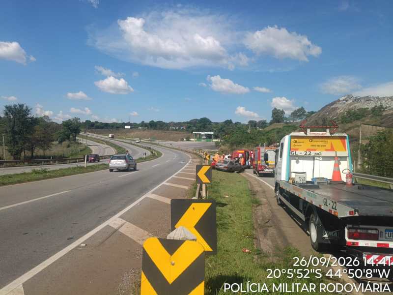 Vista de uma rodovia com carros em movimento, caminhões estacionados no acostamento e céu azul com nuvens.