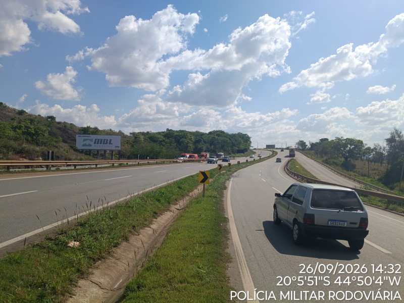 Estrada com veículos em movimento sob céu azul com nuvens, incluindo um carro cinza e um outdoor ao fundo.