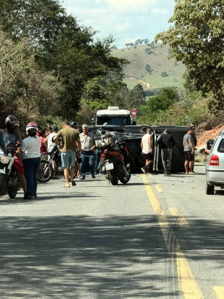 Grupo de pessoas e motociclistas em uma estrada, com um veículo tombado ao fundo.