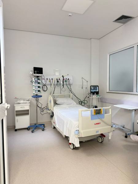 Quarto de hospital com cama vazia, equipamentos médicos ao fundo e um armário ao lado.