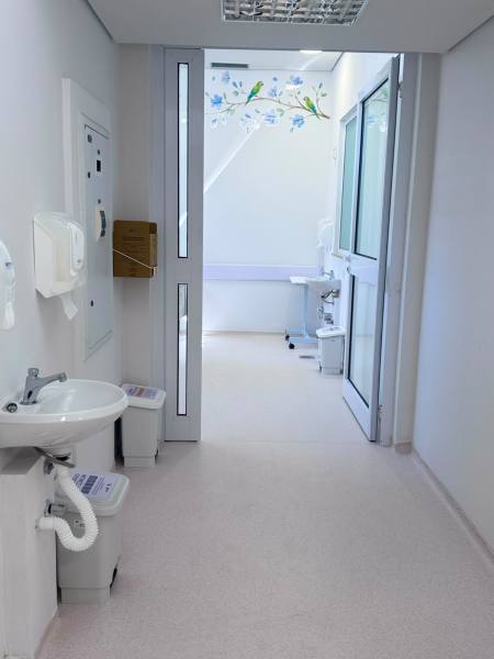 Corredor de um consultório médico com paredes brancas, contendo uma pia e dispensadores de papel toalha. Ao fundo, há uma mesa de exame e detalhes decorativos de flores e pássaros.
