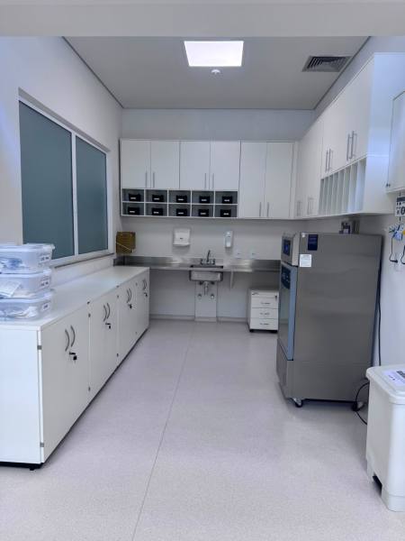 Imagem de um ambiente de cozinha ou laboratório limpo, com armários brancos, prateleiras e uma pia ao centro, além de uma geladeira cinza à direita.