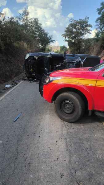 Veículo deitado na estrada após um acidente, com caminhonete dos bombeiros em primeiro plano.