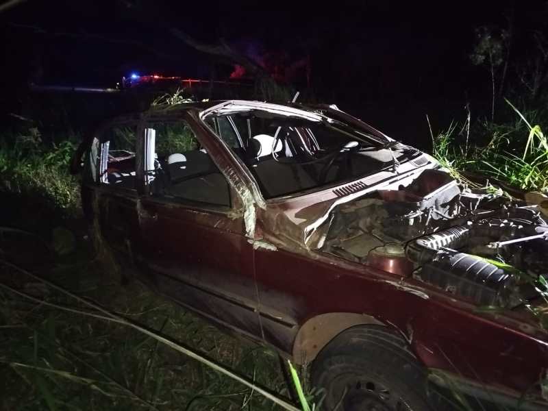 Um carro antigo e danificado, sem telhado, parado em uma área de vegetação à noite, com luzes vermelhas ao fundo.