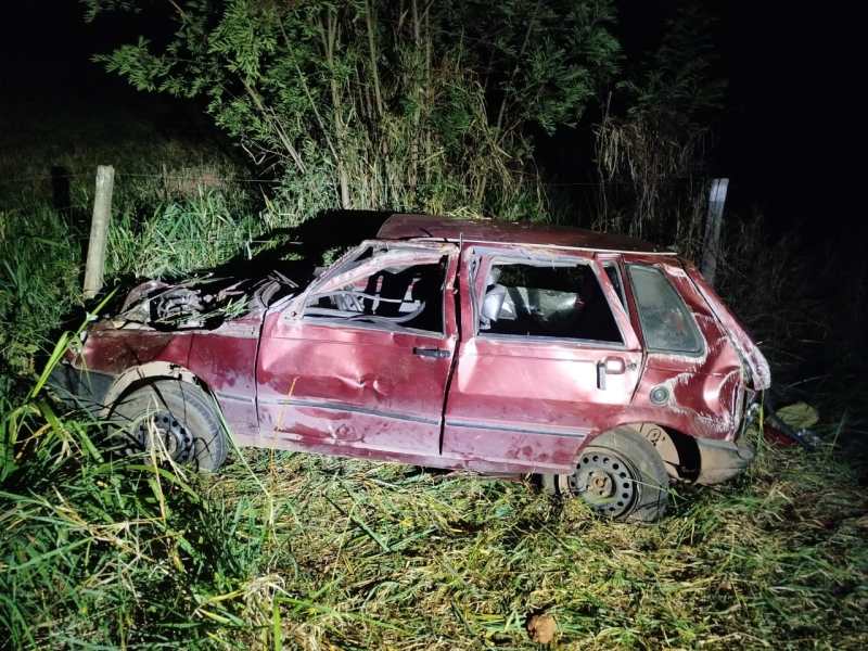 Carro acidentado e abandonado, de cor vermelha, com danos visíveis, cercado por vegetação à noite.