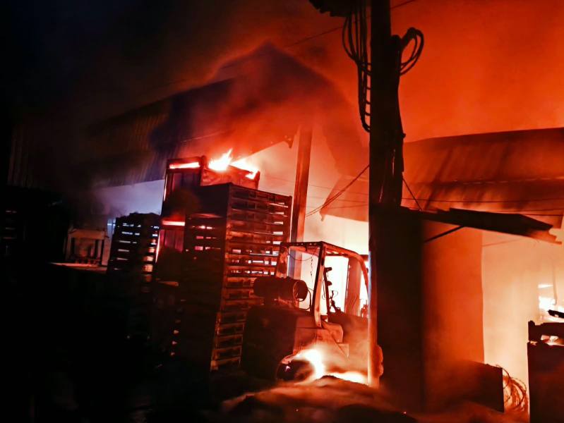 Um armazém em chamas, com fumaça e fogo visíveis. Estruturas de madeira e uma empilhadeira são parcialmente iluminadas pelo incêndio.