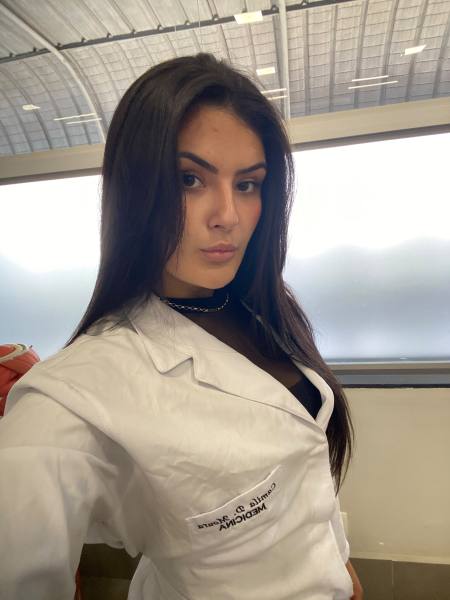 A person wearing a white lab coat with a medical logo, striking a confident pose in an indoor setting.