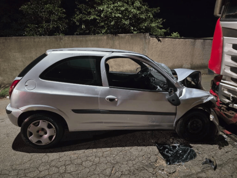 Carro prateado colidido com um caminhão à noite, com danos visíveis na lateral do veículo.