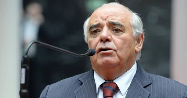 Homem mais velho falando em um microfone, usando paletó e gravata em ambiente formal.