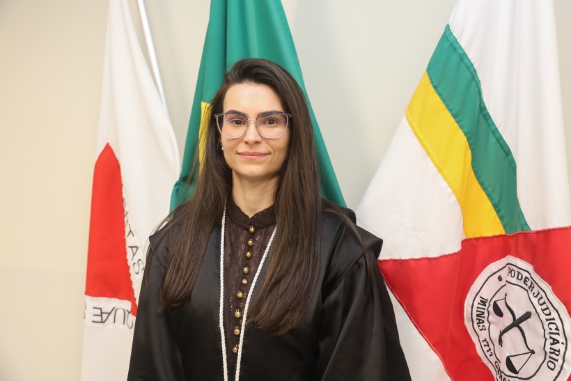Pessoa vestindo uma toga e óculos, sorrindo, em frente a bandeiras do Brasil e de Minas Gerais.